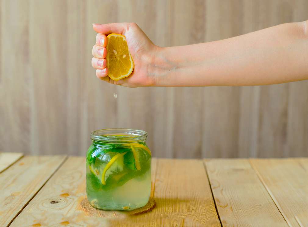 Изображение на ръка, която изстисква половин жълт лимон над чаша с лимонада