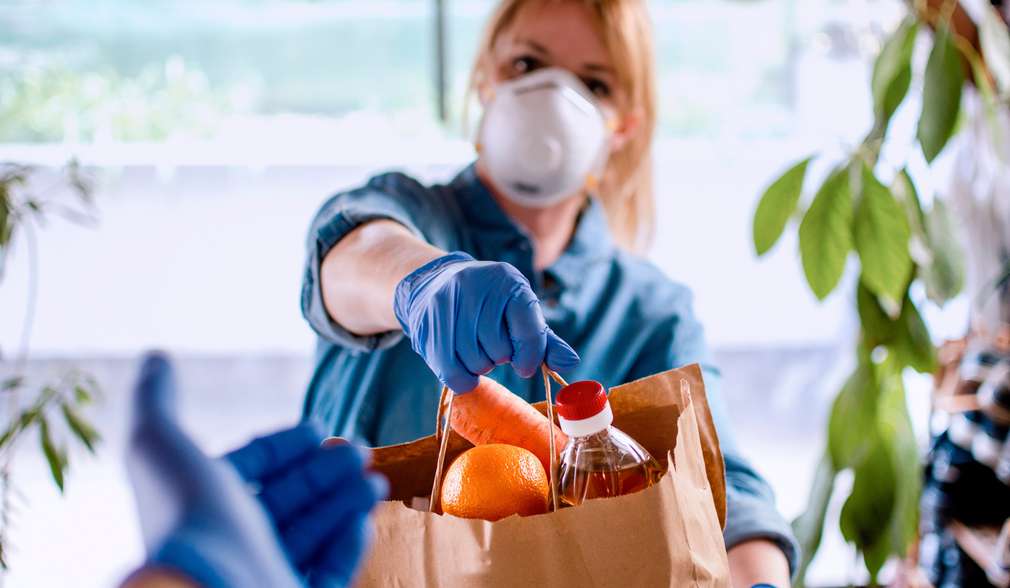 Frau mit Maske überreicht eine gefüllte Einkaufstasche aus Papier Frau mit Maske überreicht eine gefüllte Einkaufstasche aus Papier