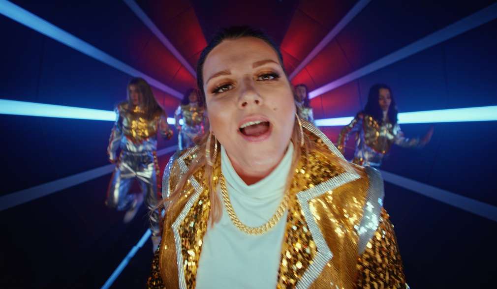 Szene aus der Online-Kampagne: Kundin im goldenen Pailetten-Blazer und Goldkette mit Tänzerinnen im Hintergrund Szene aus der Online-Kampagne: Kundin im goldenen Pailetten-Blazer und Goldkette mit Tänzerinnen im Hintergrund