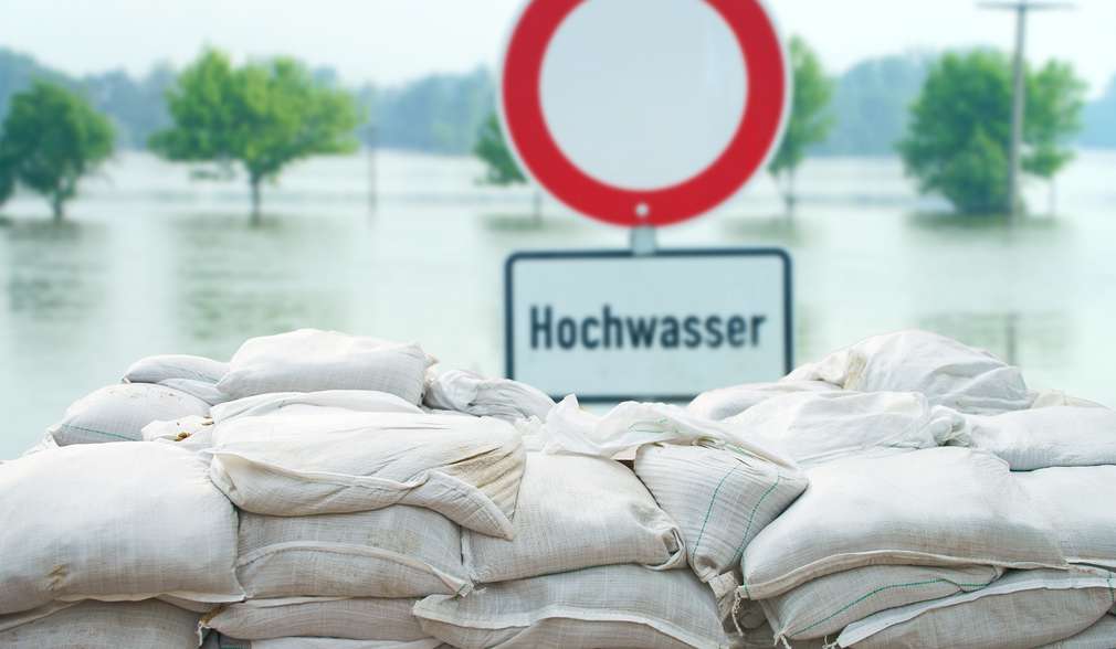 gestapelte Sandsäcke, dahinter eine überschwemmte Fläche und ein Schild „Achtung Hochwasser“ gestapelte Sandsäcke, dahinter eine überschwemmte Fläche und ein Schild „Achtung Hochwasser“