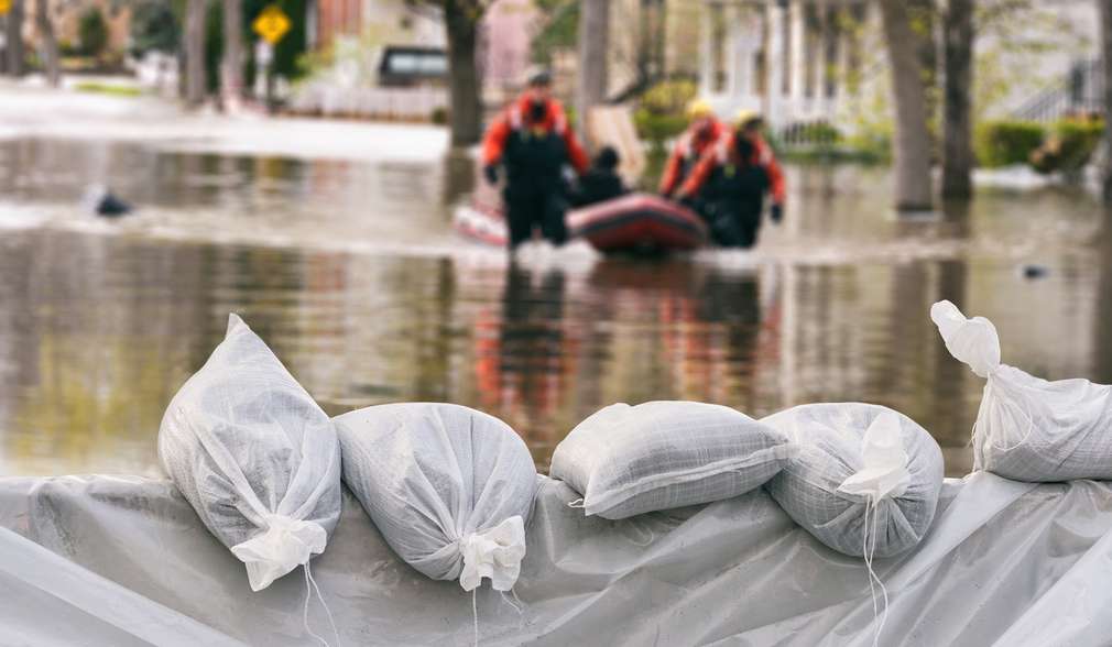 Staumauer aus Säcken gegen Hochwasser, im Hintergrund Rettungskräfte, die eine Person aus dem Hochwasser bergen Staumauer aus Säcken gegen Hochwasser, im Hintergrund Rettungskräfte, die eine Person aus dem Hochwasser bergen