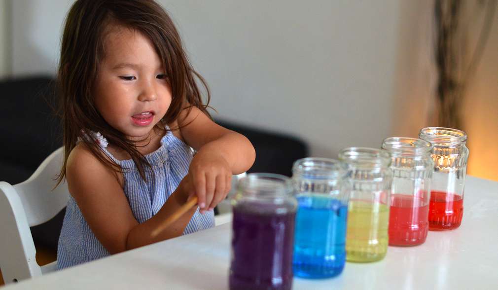 Kleines Mädchen sitzt am Tisch, darauf stehen Gläser mit bunten Flüssigkeiten befüllt Kleines Mädchen sitzt am Tisch, darauf stehen Gläser mit bunten Flüssigkeiten befüllt