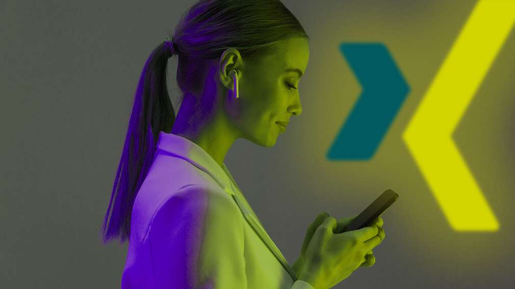 Frau im Blazer schaut auf Smartphone, hinter ihr eine Wand mit XING-Icon Frau im Blazer schaut auf Smartphone, hinter ihr eine Wand mit XING-Icon